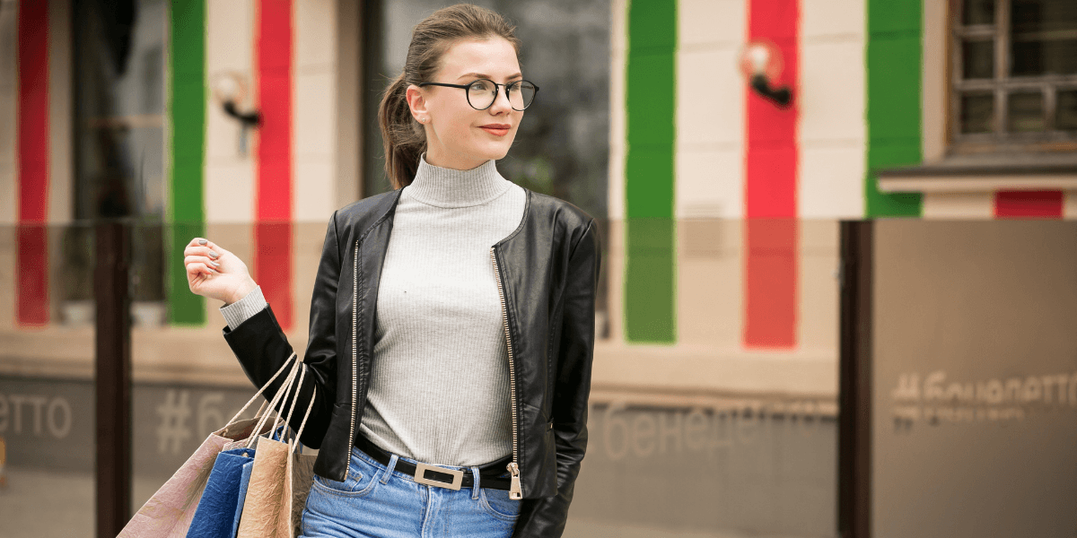 A woman in glasses, wearing a black jacket and jeans, holds shopping bags. She stands confidently against a building with vertical red and green stripes.