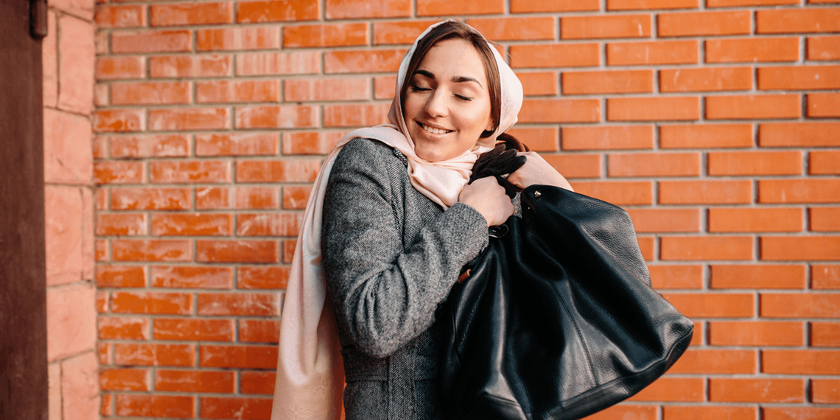Woman in a gray coat and headscarf smiles with eyes closed, clutching a large black bag, standing against a brick wall. Warm, content mood.